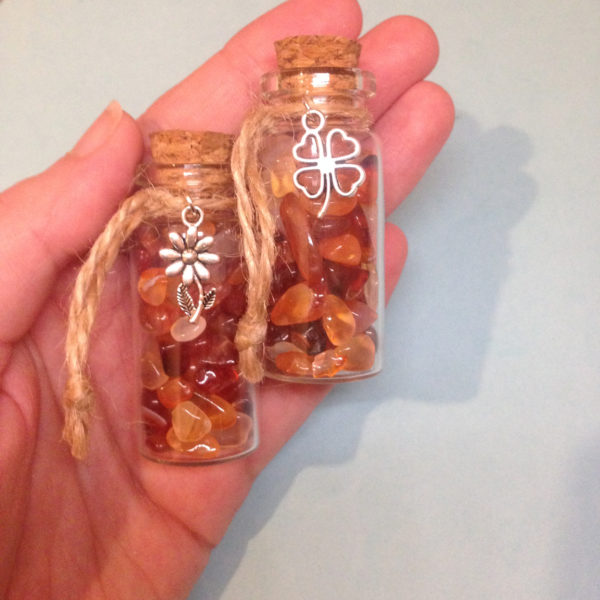 A glass jar with a cork, containing carnelian crystal chips, with a charm on twine around the top of the bottle